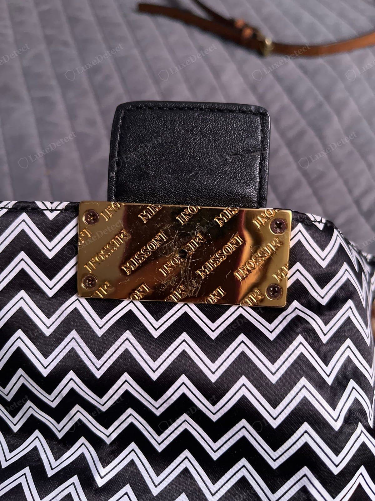 Missoni falso: el zigzag está estampado plano sobre un nailon satinado barato, con contornos blancos duros (sin textura de punto, reverso en blanco), rematado con una placa dorada rayada con el logotipo "MISSONI" repetido, atornillada al panel