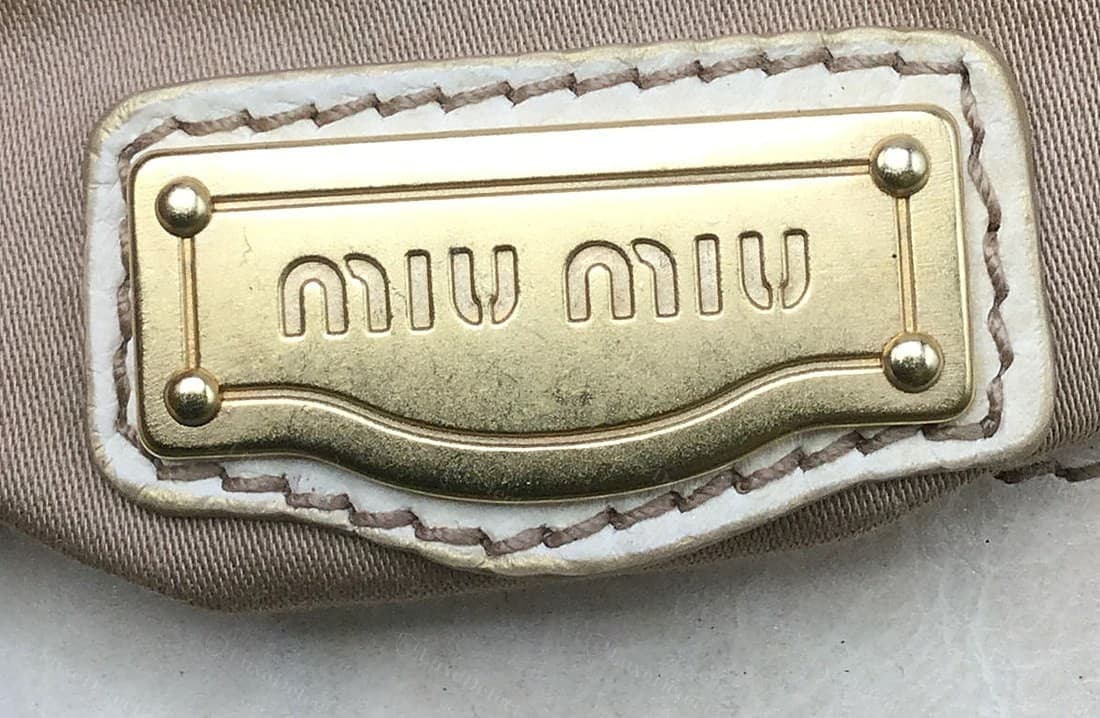 Echte Logo-Platte im Innenfutter einer Miu Miu: eine geformte Metallplakette mit eingraviertem doppeltem Rand und vier Kugelnieten, „miu miu“ in eckigen Kleinbuchstaben geprägt, auf einem Lederflicken ins Futter genäht