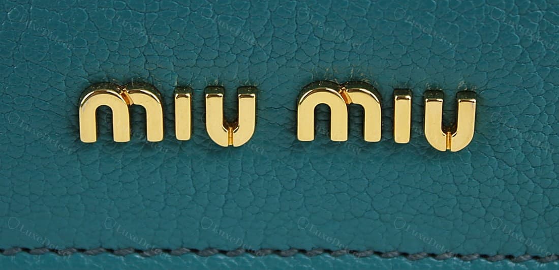 Logo exterior de un Miu Miu original: "miu miu" dos veces en letras metálicas macizas en relieve, una minúscula ancha y abierta con un grosor de trazo parejo, altura y espaciado de letras constantes, fijado con limpieza a la piel