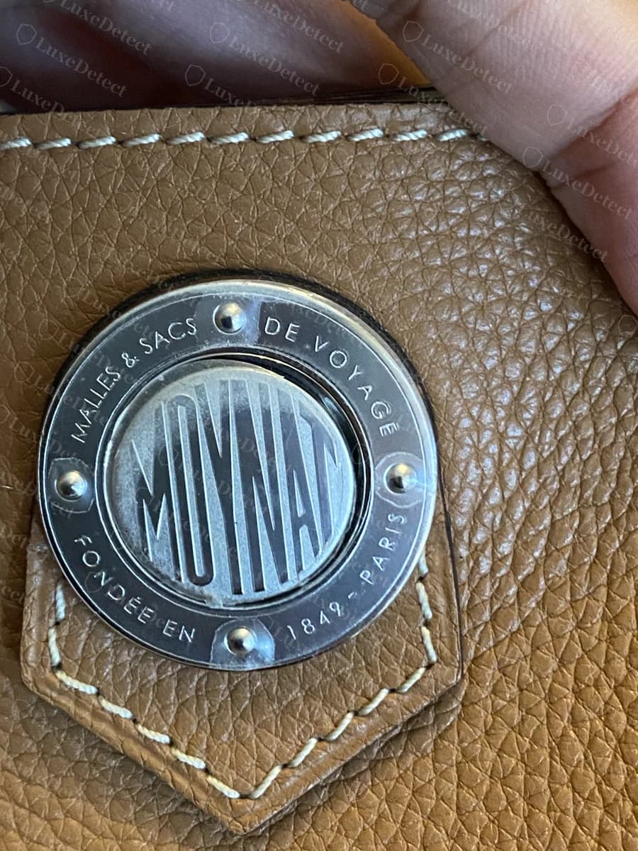 Genuine Moynat silver-tone medallion reading MALLES & SACS DE VOYAGE, FONDÉE EN 1849, PARIS around a condensed MOYNAT, on a tan leather bucket bag