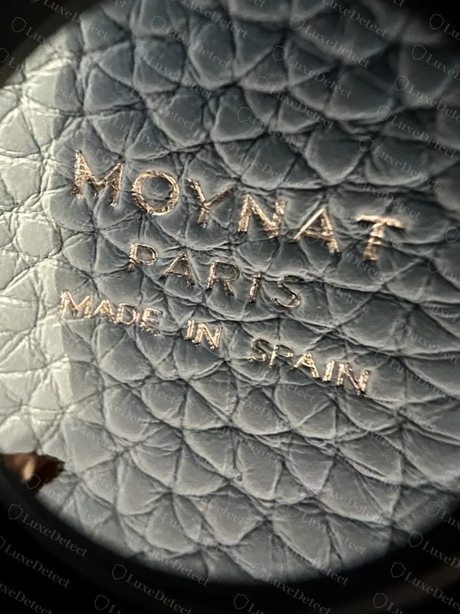 Genuine Moynat interior stamp reading MOYNAT PARIS MADE IN SPAIN, photographed under magnification on pebbled blue leather