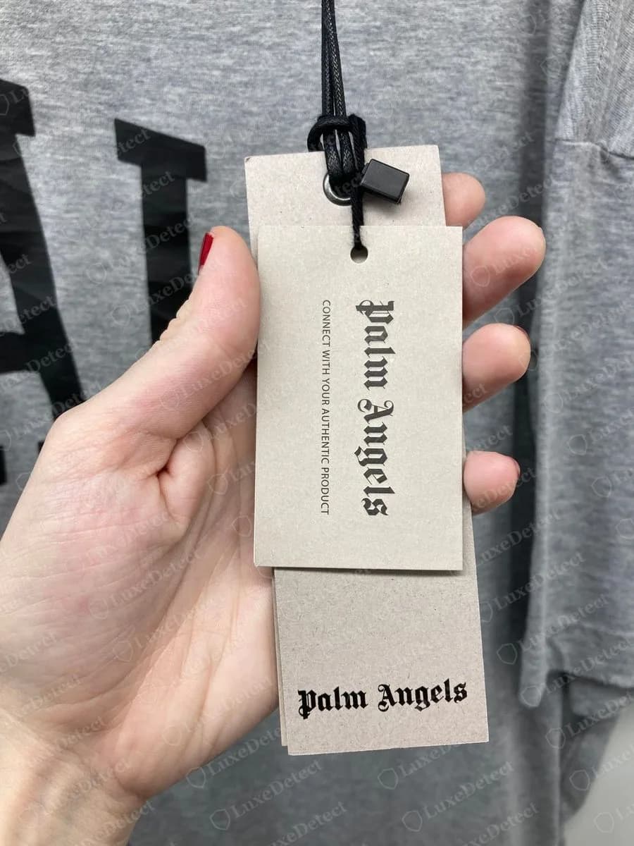 Genuine Palm Angels grey card hangtag reading CONNECT WITH YOUR AUTHENTIC PRODUCT in gothic lettering, hanging on a cord from a grey T-shirt