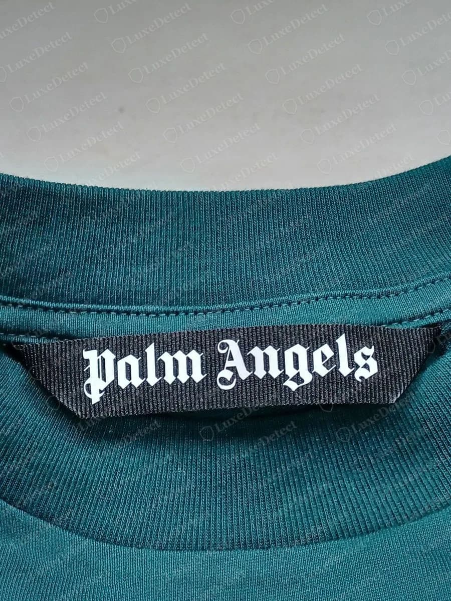 Genuine Palm Angels black woven neck label with the name in white gothic lettering, sewn at the inside back neck of a green T-shirt