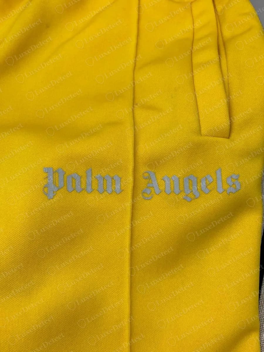 Genuine Palm Angels yellow track pants with a reflective silver-grey gothic Palm Angels logo printed across the front seam near the pocket
