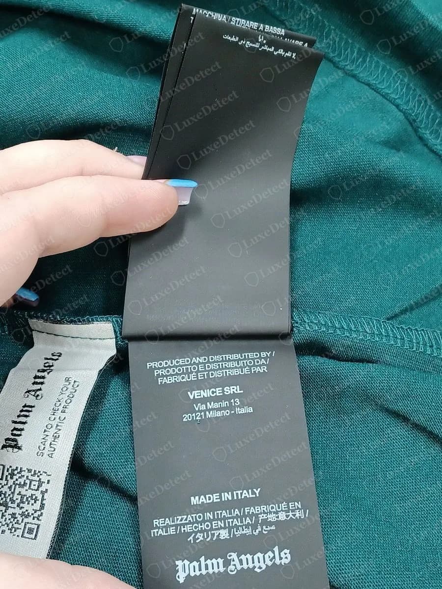 Genuine Palm Angels black wash tag showing PRODUCED AND DISTRIBUTED BY, VENICE SRL, a Milan address and MADE IN ITALY, beside a grey woven QR label
