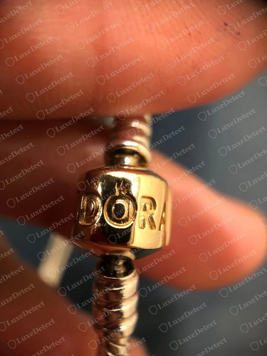Echter goldfarbener Pandora Tonnenverschluss eines zweifarbigen Armbands, mit DORA und einer Krone über dem O, zwischen Fingern gehalten