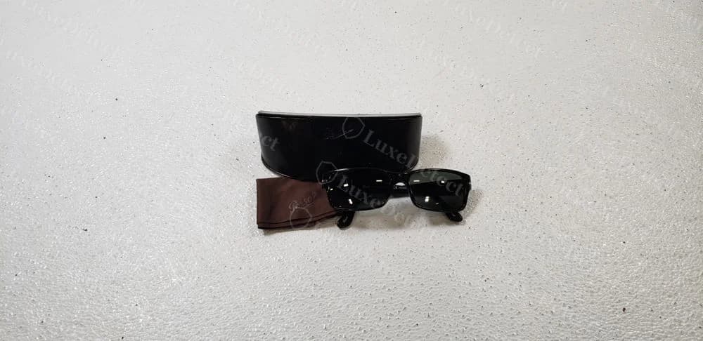 Gafas de sol Persol negras originales junto a un estuche rígido negro con el logotipo Persol en cursiva en la tapa y un paño marrón estampado con la cursiva