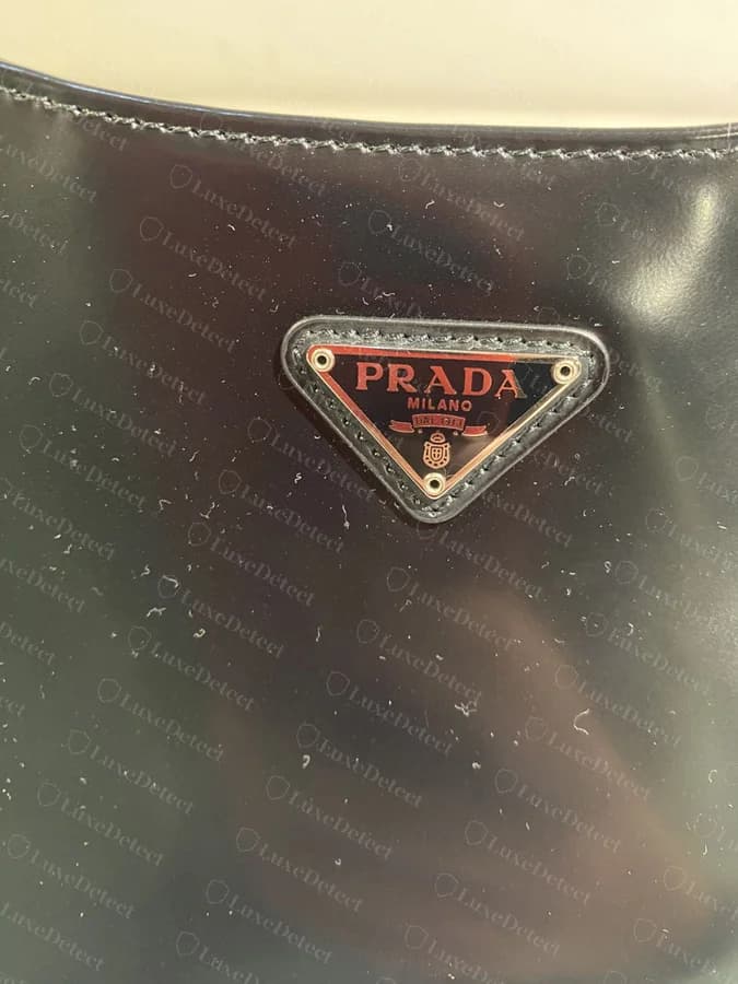 Counterfeit Cleo triangle plaque: a red/burgundy tint visible under the PRADA MILANO lettering and ribbon border