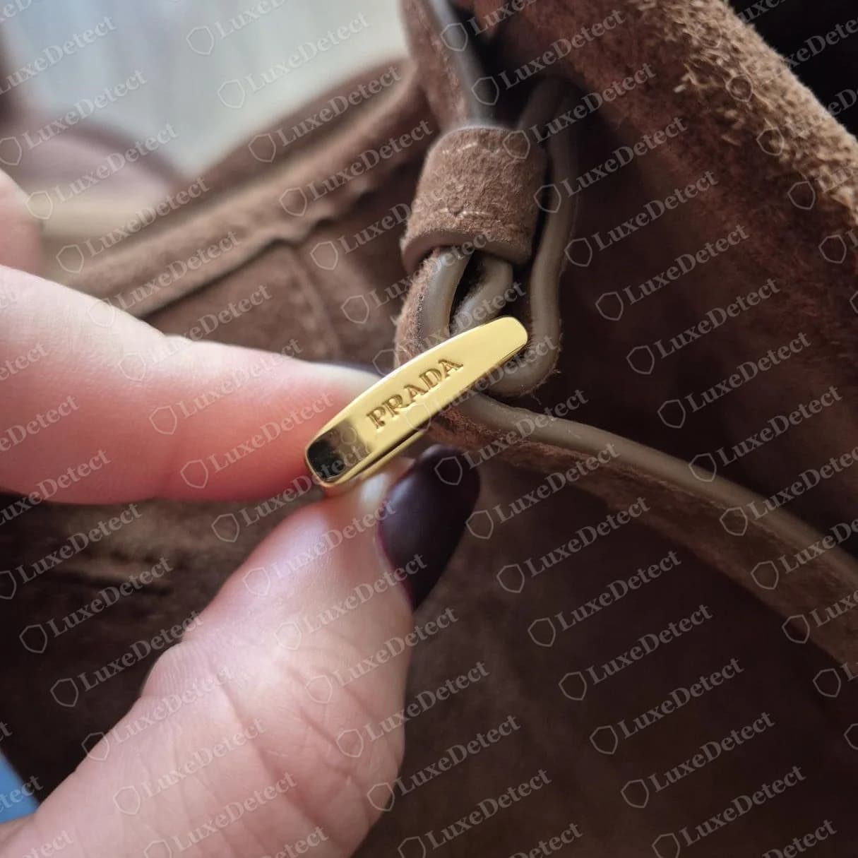 Bolsa de ombro de camurça Prada falsa: puxador dourado em forma de barra com “PRADA” gravado com capricho e centralizado. A gravação parece correta, por isso não entrega essa falsificação