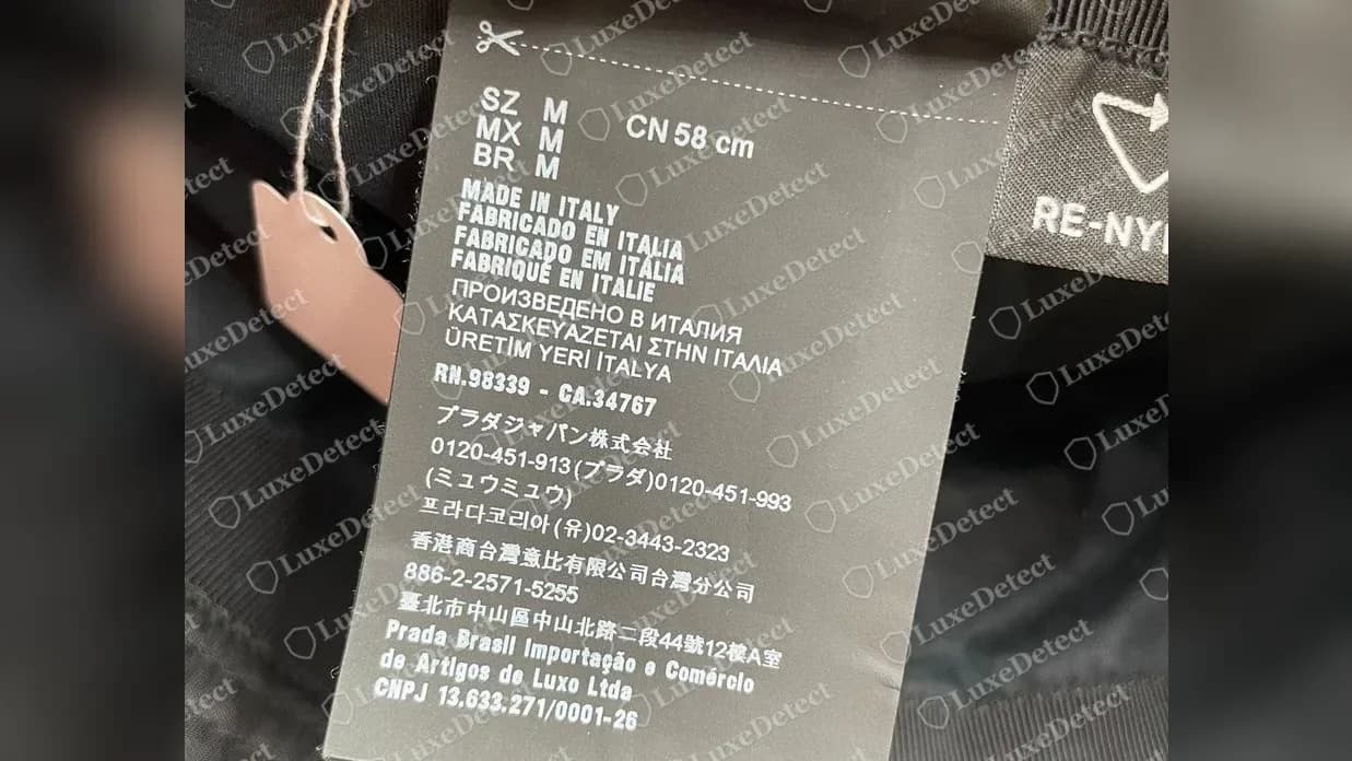 Etichetta multilingue di un bucket hat Prada originale: taglia M, CN 58 cm, MADE IN ITALY in diverse lingue, RN.98339 - CA.34767 e gli indirizzi di importazione di Prada