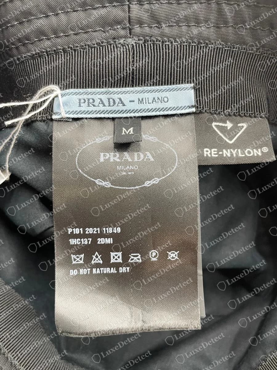 Genuine Prada Re-Nylon bucket hat labels: the pale blue PRADA - MILANO ribbon, a black logo label with a size M tab and the codes P101 2021 11849 and 1HC137 2DMI, and the RE-NYLON tag beside it