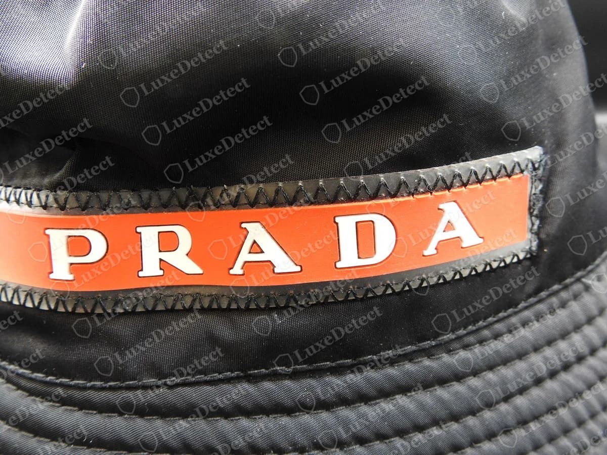 Gorro bucket hat Prada falso con una banda PRADA roja cosida en el frente en lugar de la placa triangular; por dentro llevaba la misma etiqueta de lavado ACU1 en inglés que las demás falsificaciones