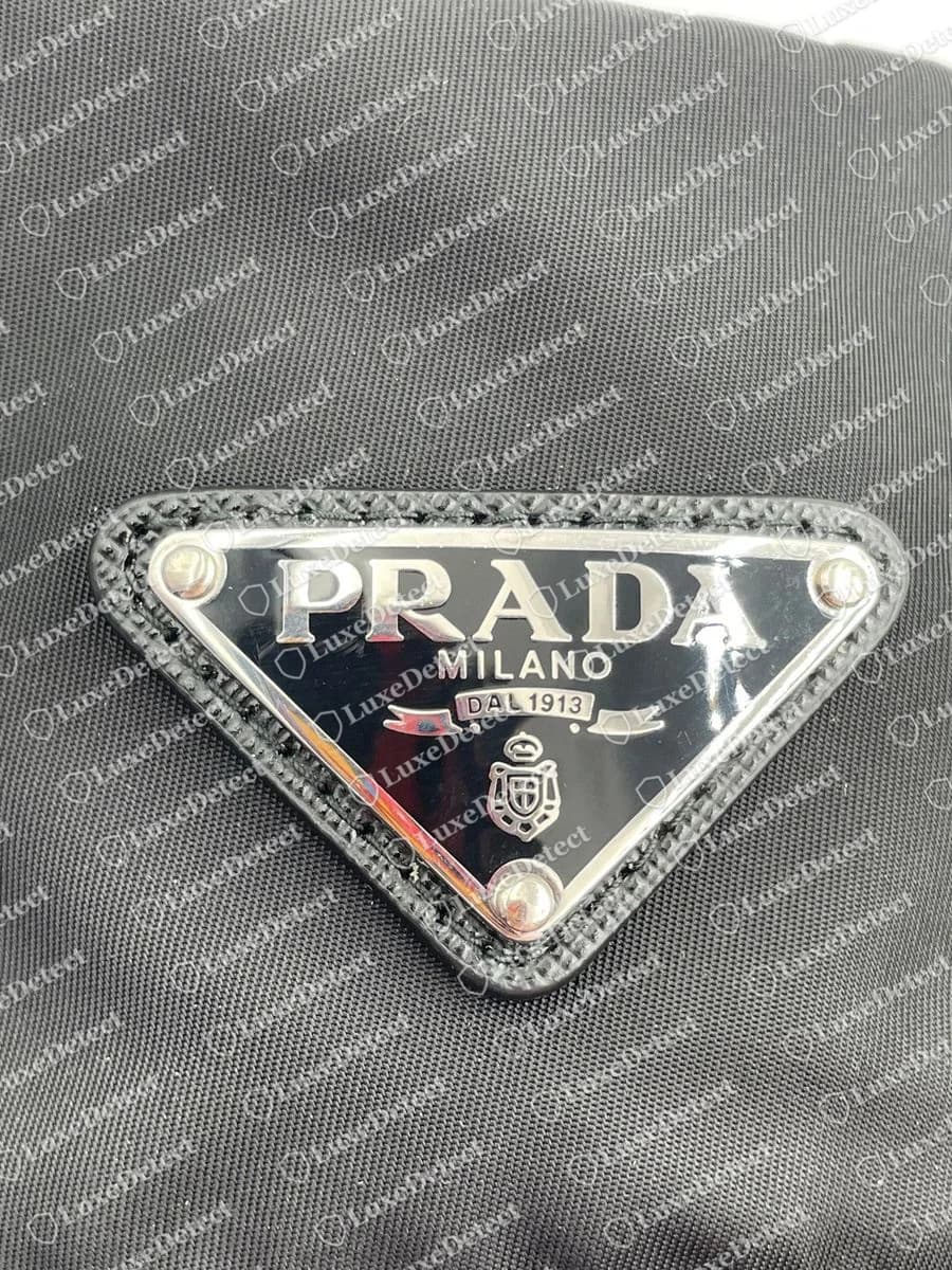 Placca a triangolo di un bucket hat Prada originale: metallo lucido color argento con smalto nero, lettere PRADA e MILANO in rilievo, DAL 1913 su un nastro, uno stemma e tre rivetti bombati su una cornice in pelle saffiano