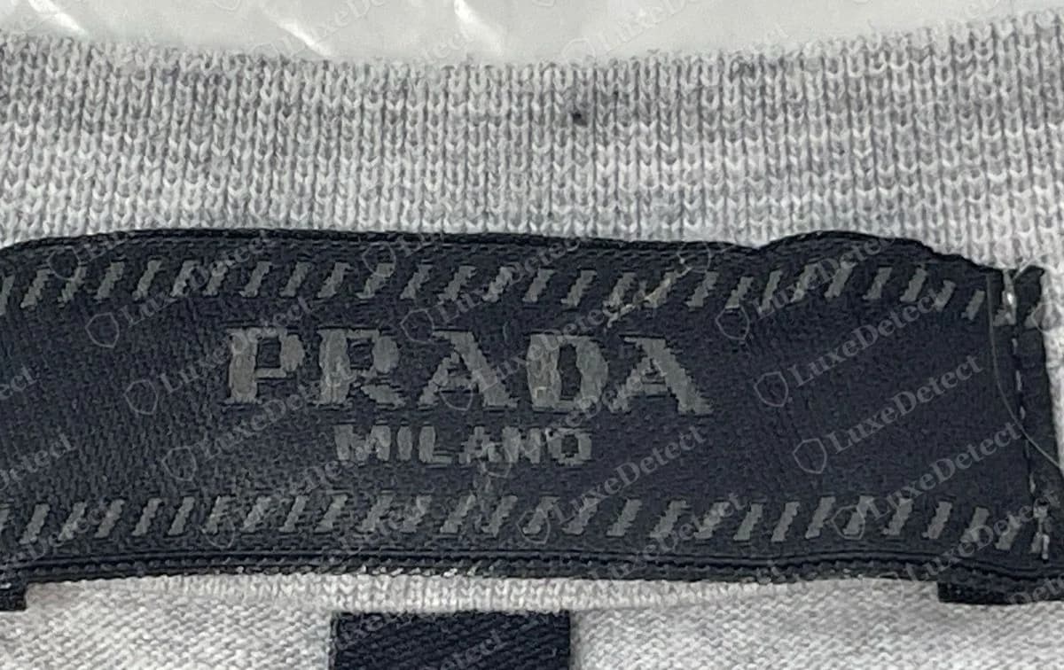 Halsetikett eines gefälschten Prada T-Shirts: ein schwarzes gewebtes Etikett „PRADA / MILANO“ mit denselben gestreiften Rändern. Solche Etiketten lassen sich mit bloßem Auge nicht von echten unterscheiden.