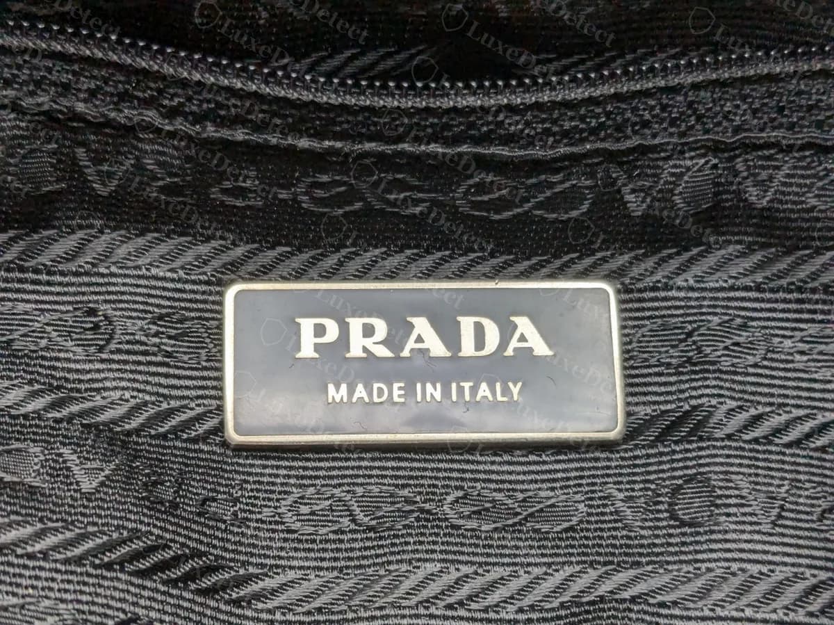 Genuine Prada backpack interior: a black rectangular plate reading PRADA and MADE IN ITALY on a woven lining patterned with repeating PRADA lettering and rope-like bands