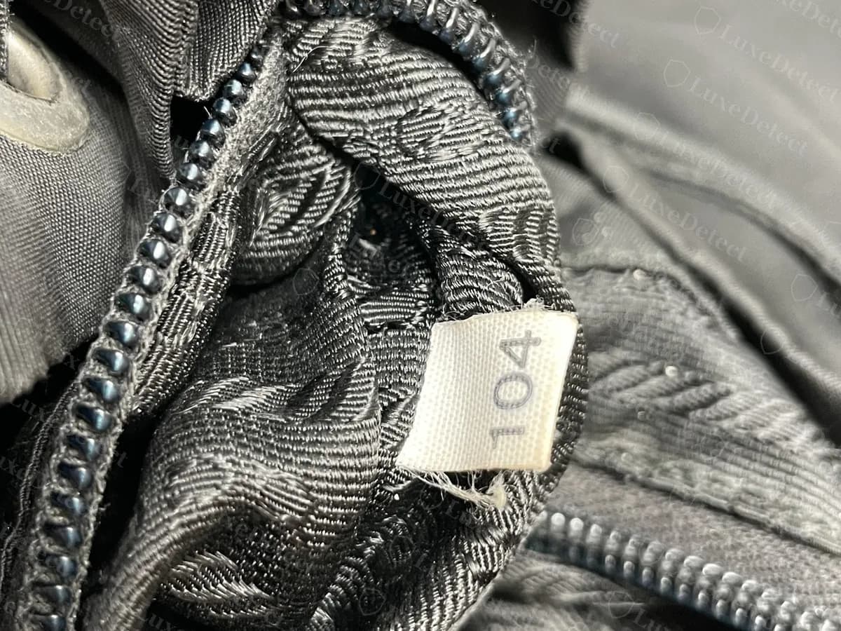Small white fabric tag with the number 104 sewn into the lining of a genuine Prada nylon backpack, beside the zip