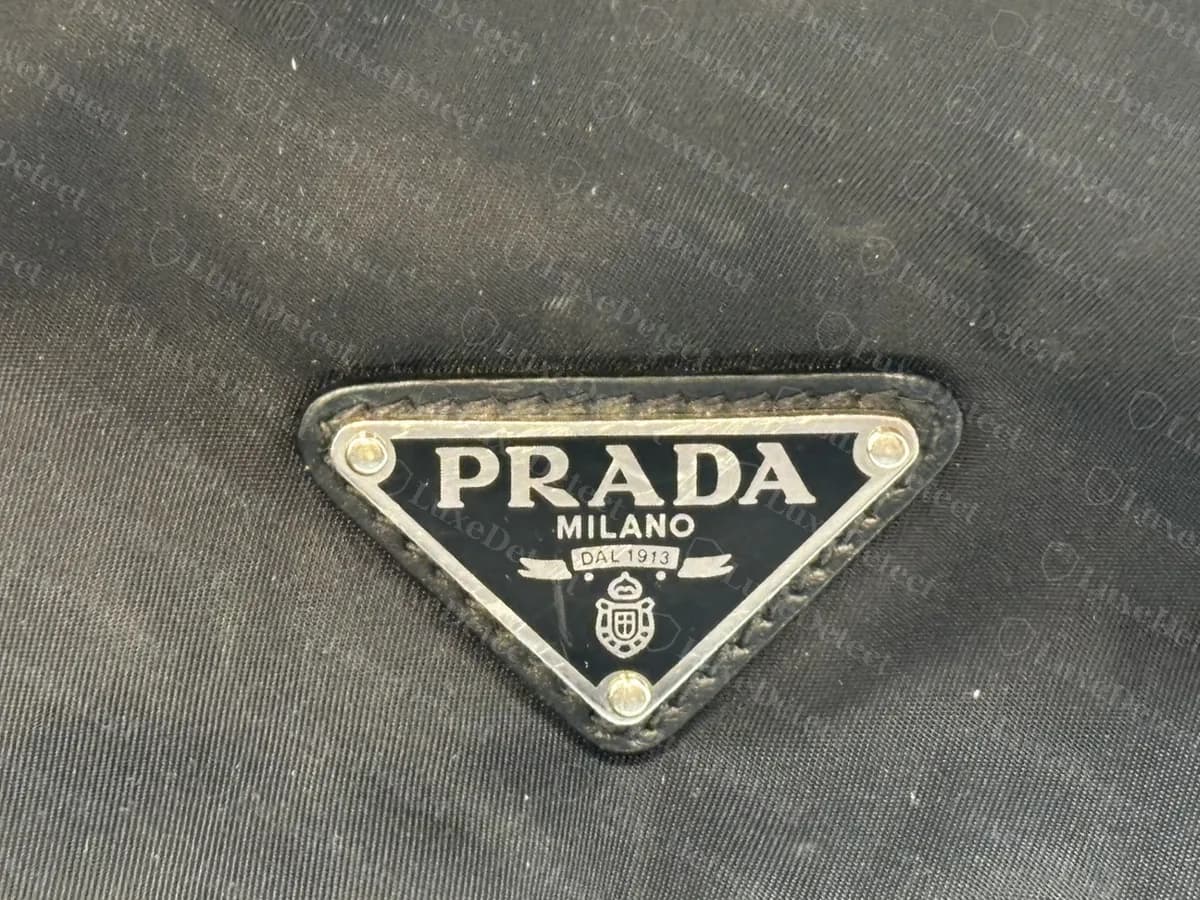 Genuine Prada nylon backpack triangle logo: a black enamel and silver-tone plate reading PRADA, MILANO and DAL 1913 above a small crest, riveted at the corners onto a leather-edged base