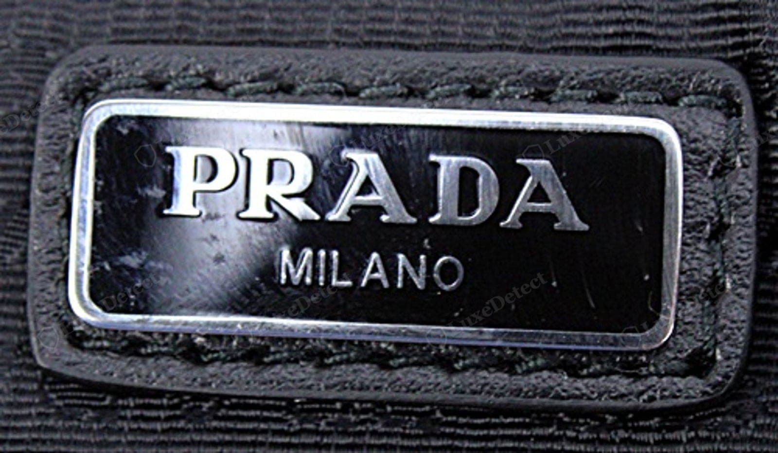 Placa triangular de metal de un Prada Re-Edition original: "PRADA" sobre "MILANO" y la línea de fundación nítidos bajo la lupa, esmalte brillante y a nivel