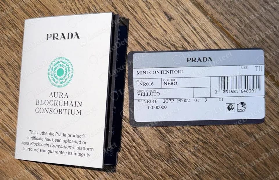 Carte Prada authentique indiquant le nom du produit, le code article, la couleur et la matière, à côté d'un dépliant de certificat Aura Blockchain Consortium d'un achat récent