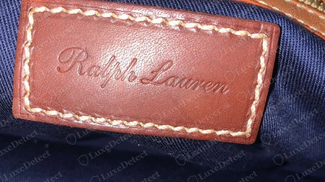 Patch interno de uma Ricky da Ralph Lauren original: um painel de couro marrom com “Ralph Lauren” em baixo-relevo em letra cursiva, contornado por pespontos cor creme, sobre forro de lona azul-marinho