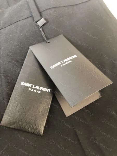 Etiquetas colgantes genuinas de Saint Laurent Paris, cartulina negra con el logotipo en mayúsculas blancas, sobre unos pantalones negros.