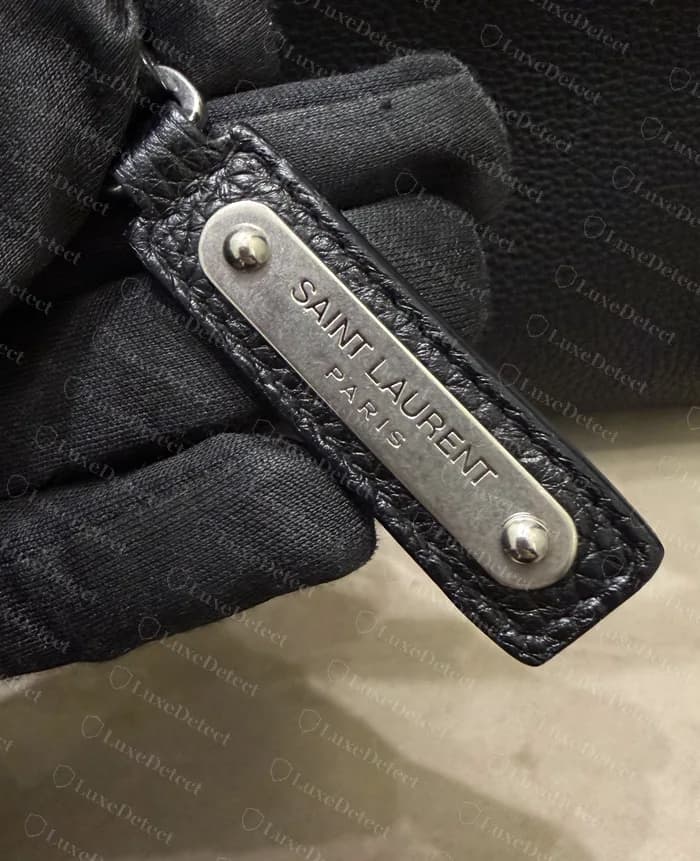 Genuine Sac de Jour name tag: grained black leather with a silver plate held by two domed rivets, stamped SAINT LAURENT over PARIS