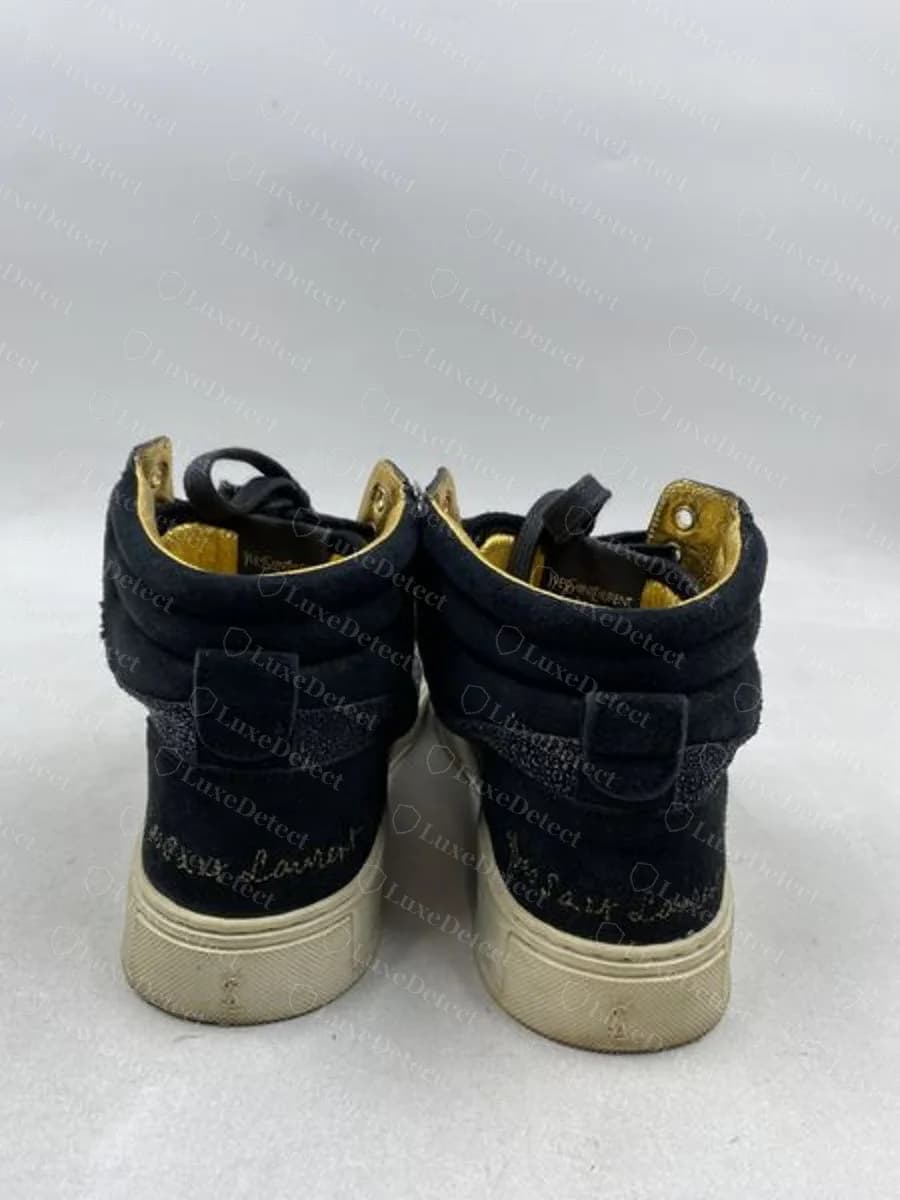 Genuine "Yves Saint Laurent" wordmark embroidered in gold thread directly into the suede heel counter of a black high-top sneaker, not printed or foil-stamped.