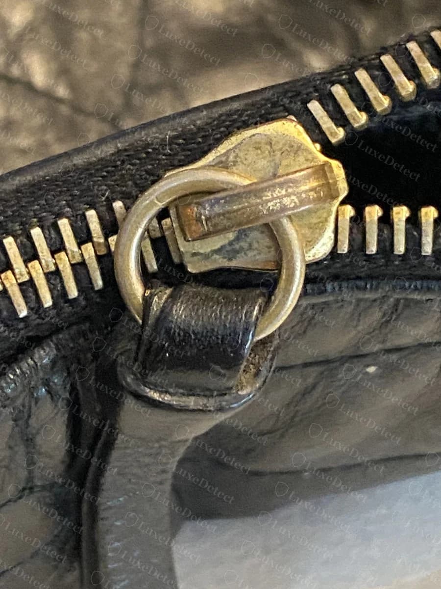 The zipper-pull hardware on the main compartment, a rounded brass tag secured through a leather zip tab.