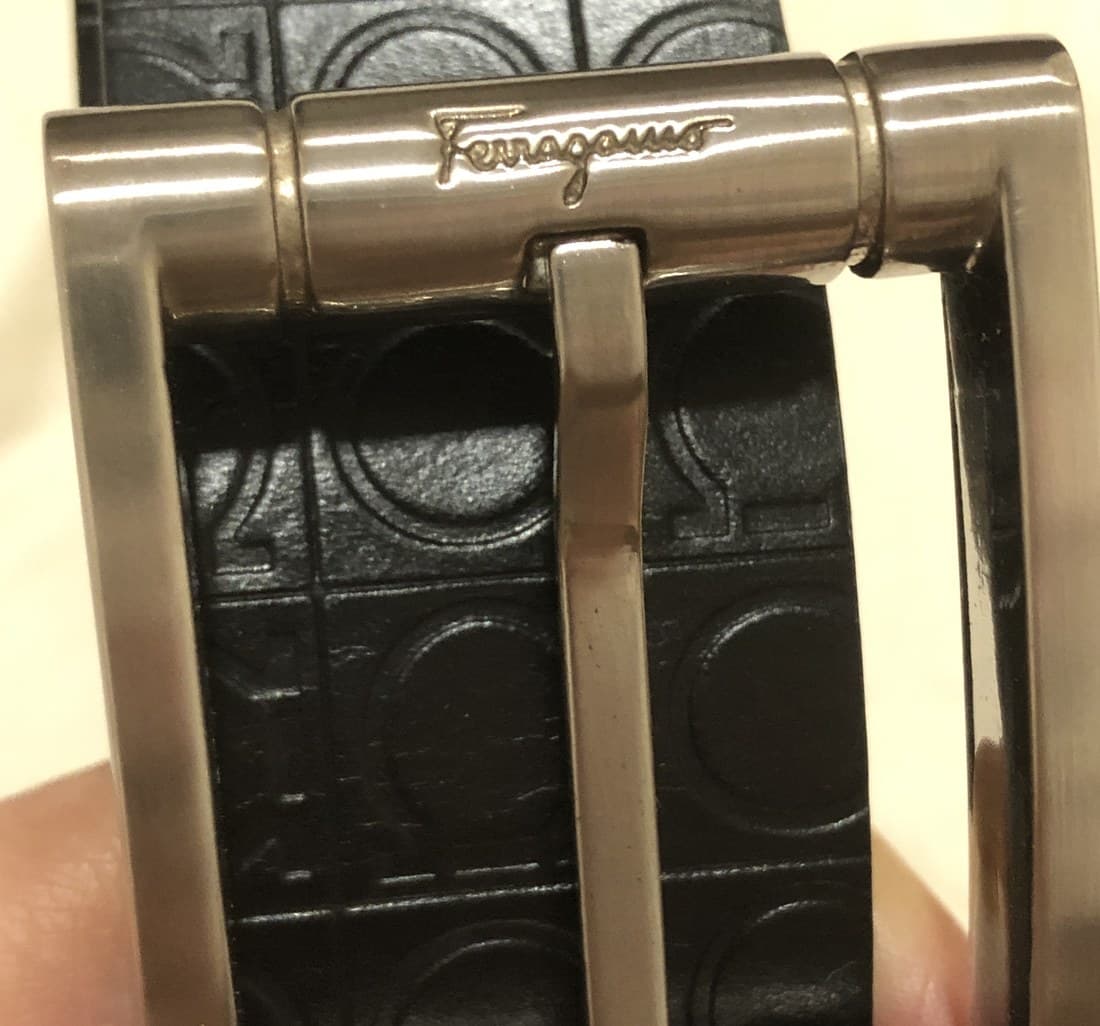 Genuine Salvatore Ferragamo Gancini buckle on a men's belt — the metal frame shows a ribbed, sunburst-textured finish radiating from the rings, with "Ferragamo" engraved in script on a separate flat polished plate