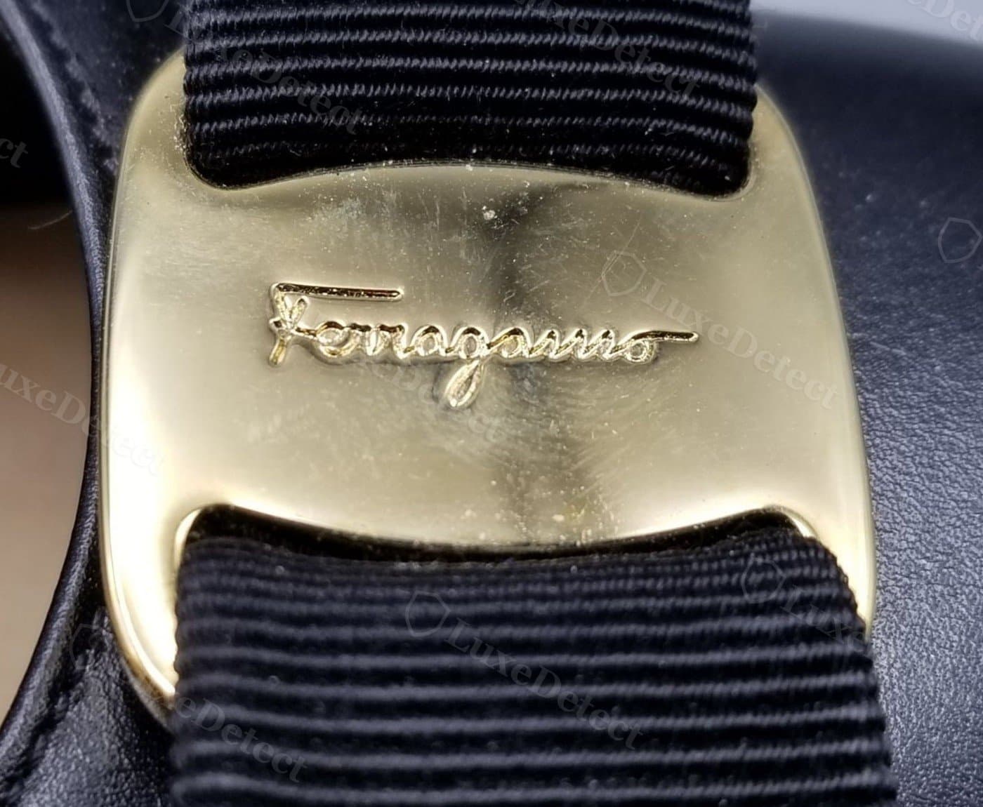 Primer plano de un cierre Vara original: la placa central dorada fundida lleva una grafía "Ferragamo" nítidamente grabada, con profundidad real, engastada en una cinta de grosgrain azul marino de tejido apretado