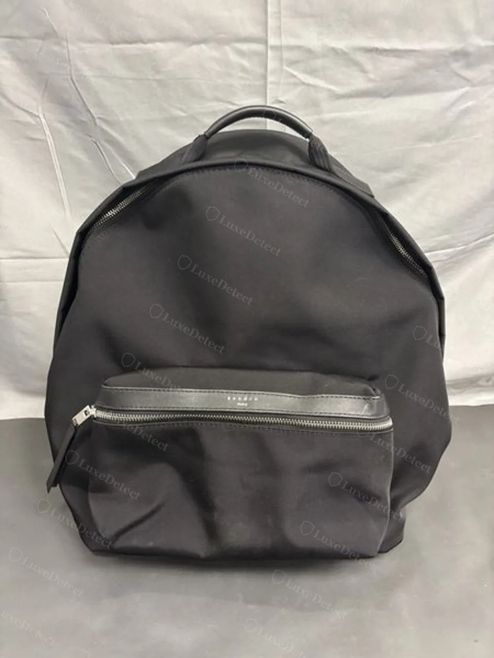 Front exterior of Sandro's black nylon book bag backpack, showing the leather-trimmed top handle and the embossed wordmark panel on the front zip pocket.