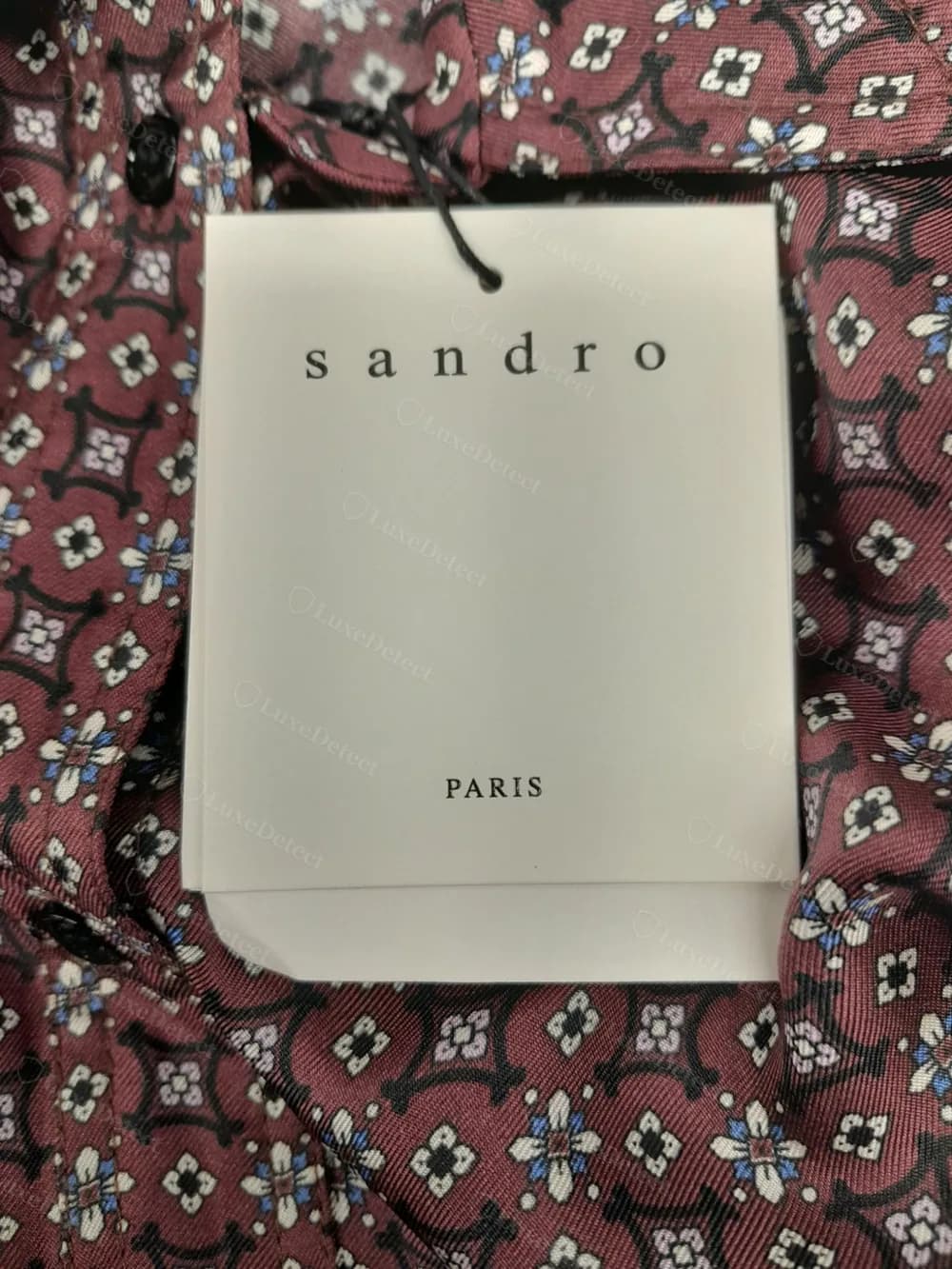 Folded cardstock "sandro / PARIS" hangtag attached by a black cotton loop, pinned to a silk print shirt.