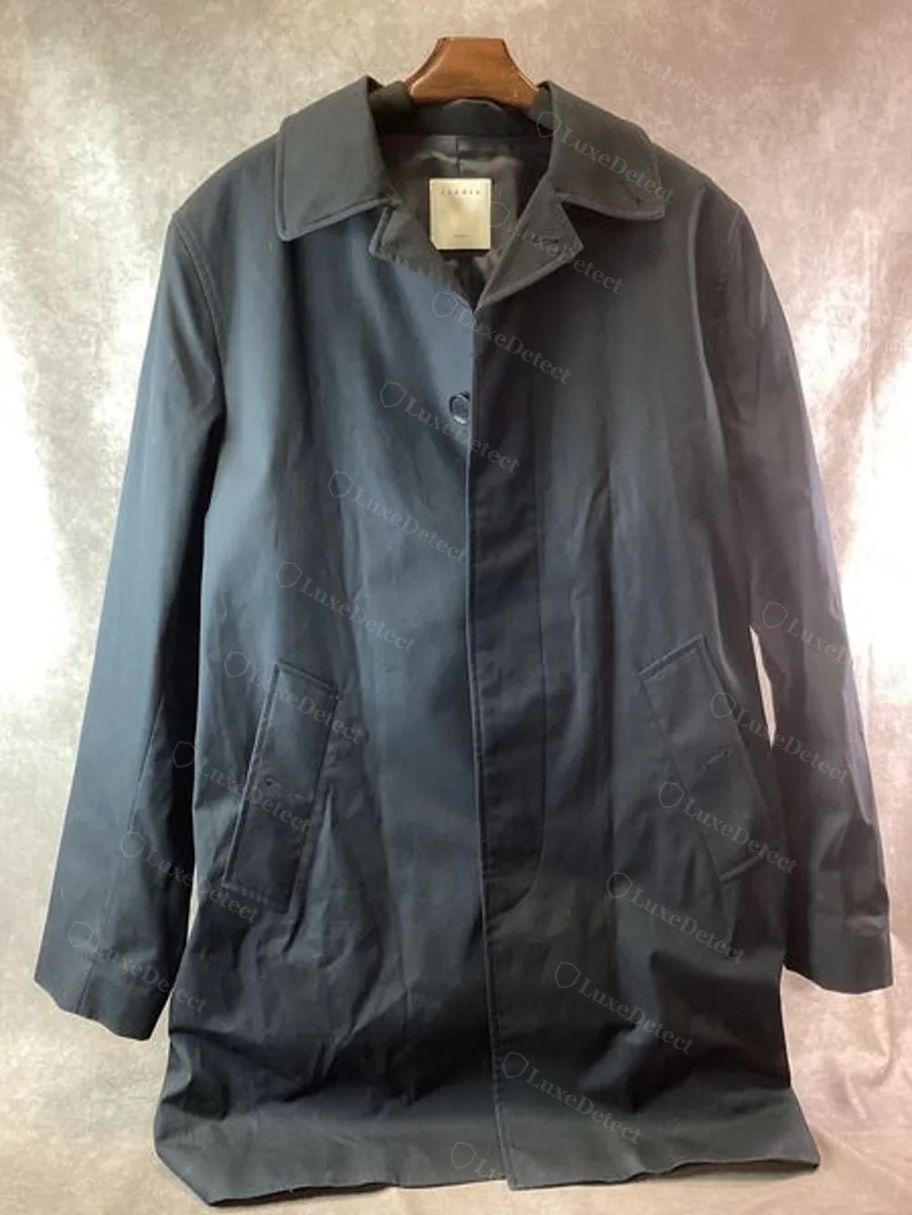 A men's cotton trench coat on a hanger, showing the front placket, welt pockets, and interior collar label positioned just below the collar seam.