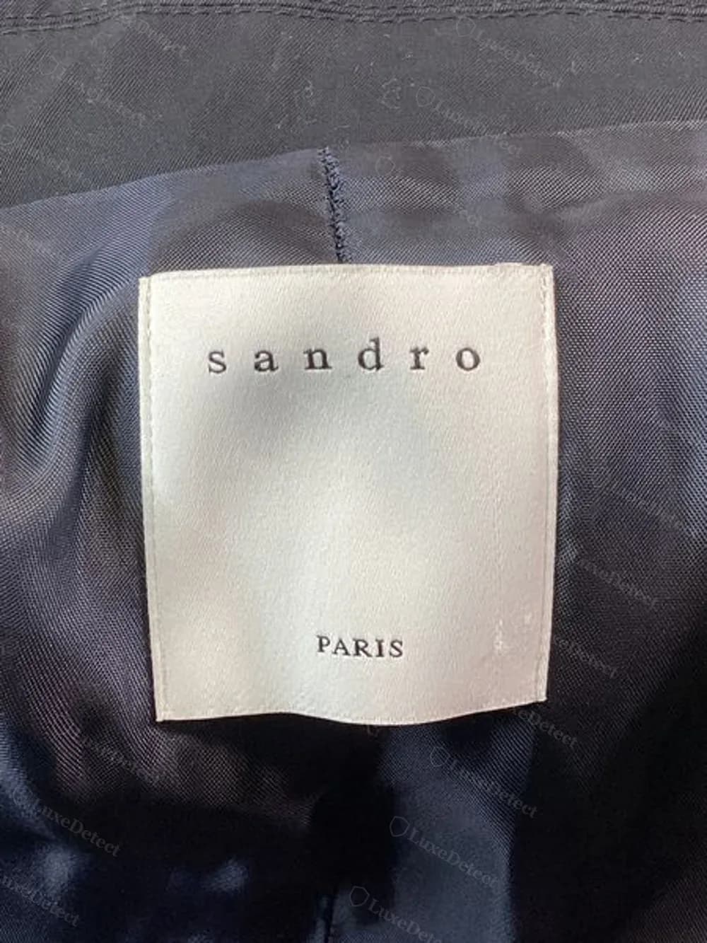 Woven "sandro PARIS" label sewn into a men's cotton trench coat, cream ground with the wordmark in dark serif type.