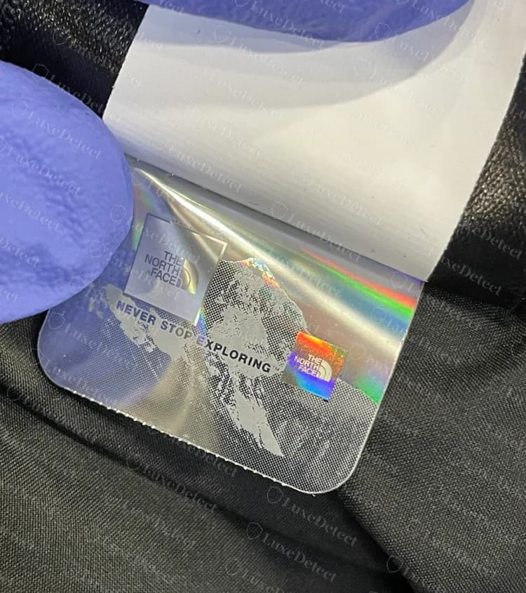 Pegatina con holograma genuina en una lengüeta doblada dentro de una parka de plumas: foil arcoíris, el logo y las palabras Never Stop Exploring