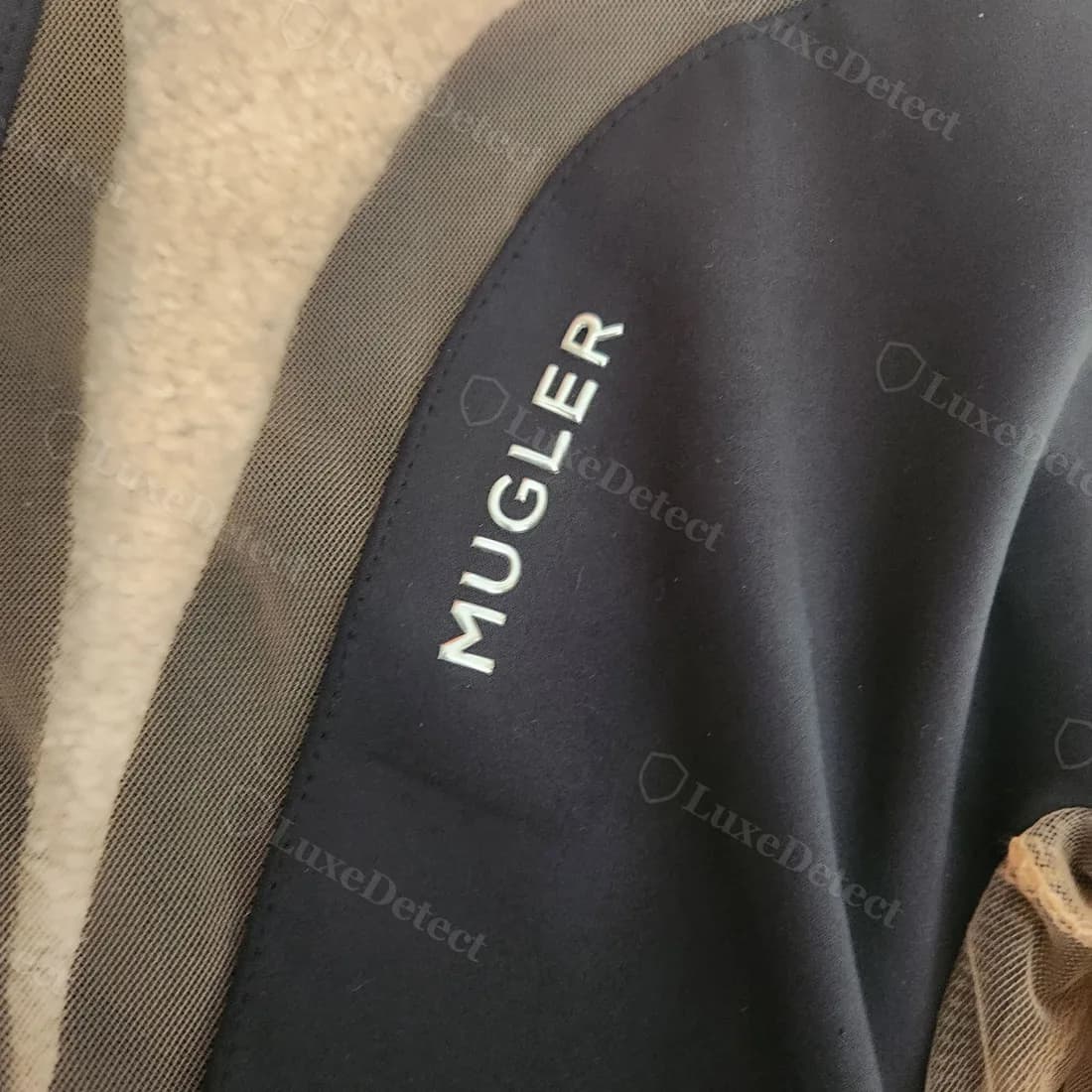 Metallic transfer-print "MUGLER" wordmark at the shoulder of a confirmed-fake illusion bodysuit: slightly raised, glossy finish rather than woven into the fabric.