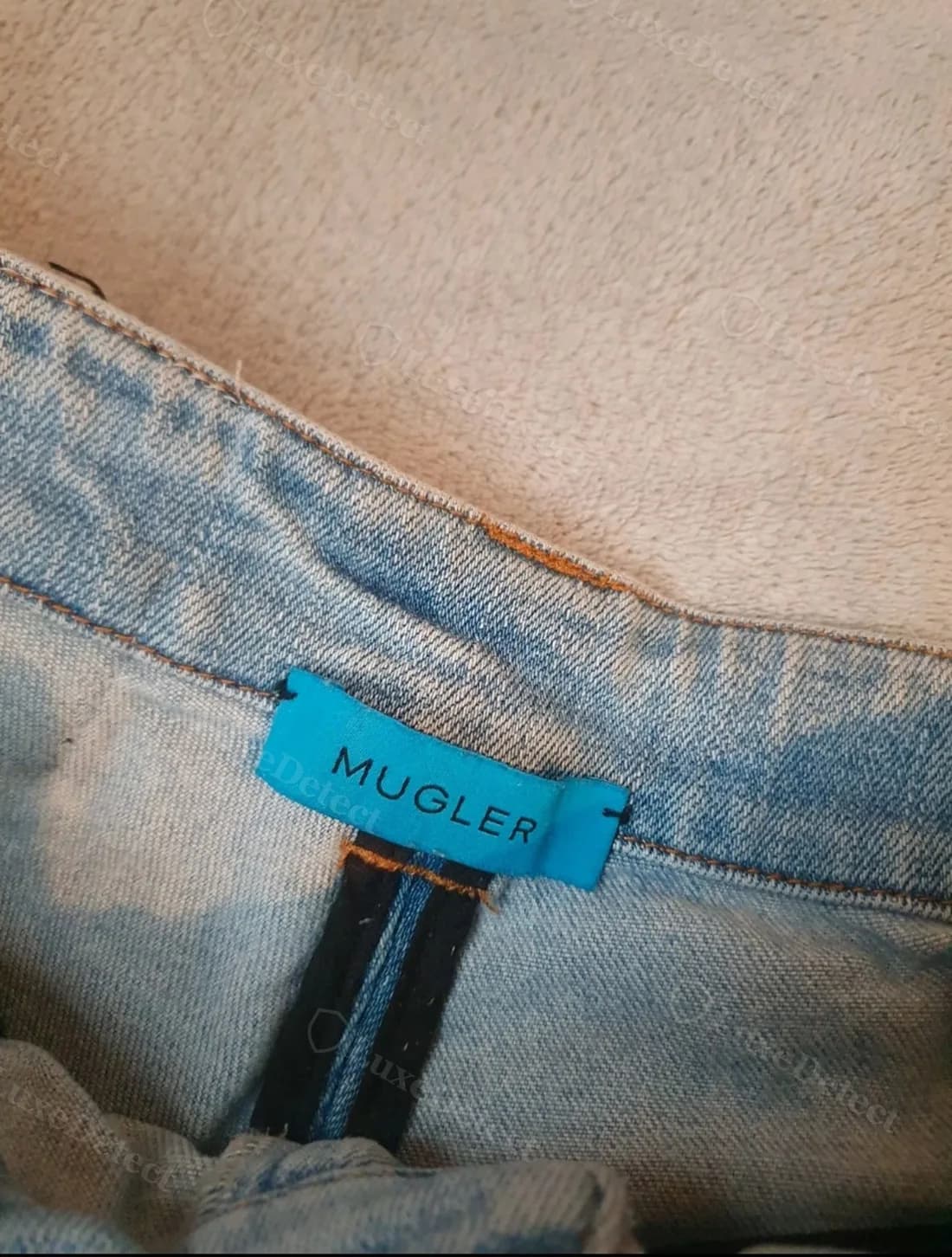 Authentic waistband tag on the same jeans: teal satin loop tag, evenly kerned "MUGLER" wordmark, tight stitching at both ends.