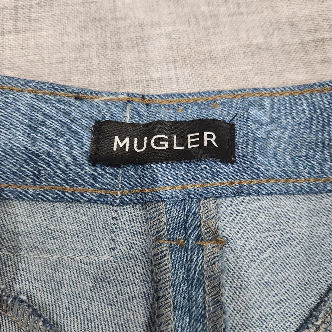 Woven "MUGLER" tag on confirmed-fake jeans: rounder tag corners and less even letter spacing than the authentic waistband tag above, on a different cut of denim (jodhpur-panel jeans, not the curved-seam silhouette).
