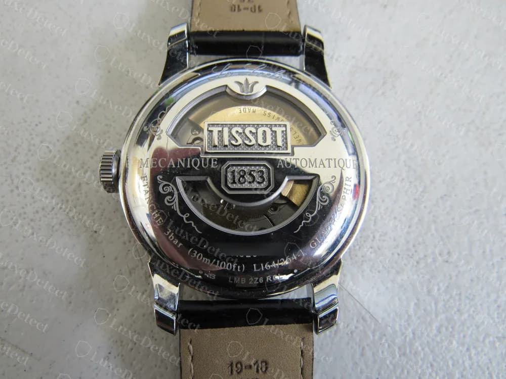 Un Tissot Le Locle Automatic original, fondo de exhibición: una placa TISSOT con 1853 en la ventana del rotor, MECANIQUE y AUTOMATIQUE a cada lado, ETANCHE 3bar (30m/100ft) y la referencia L164/264-1 en el borde. El número de serie está difuminado.