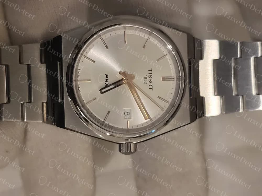 Tissot PRX original, mostrador prateado: TISSOT 1853 impresso abaixo do 12, o nome PRX em negrito mais embaixo no mostrador, índices aplicados em bastão, uma janela de calendário às 3 horas e o pequeno texto SWISS MADE perto da borda. A foto foi tirada com o relógio de lado.