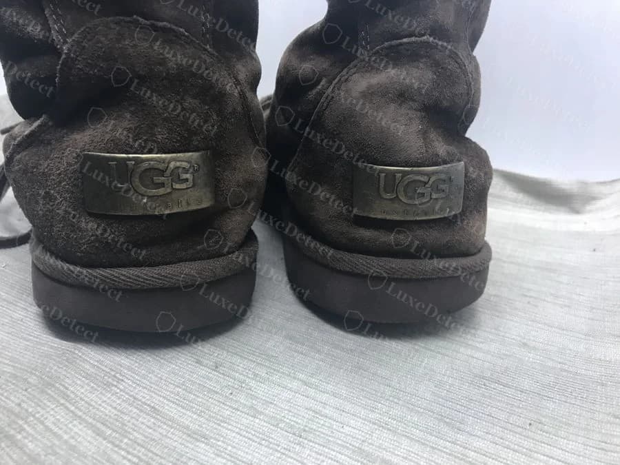 Genuine UGG metal heel plaque: a riveted brass-toned plate stamped UGG with a small registered trademark mark and australia beneath, set into the suede heel of a tall boot