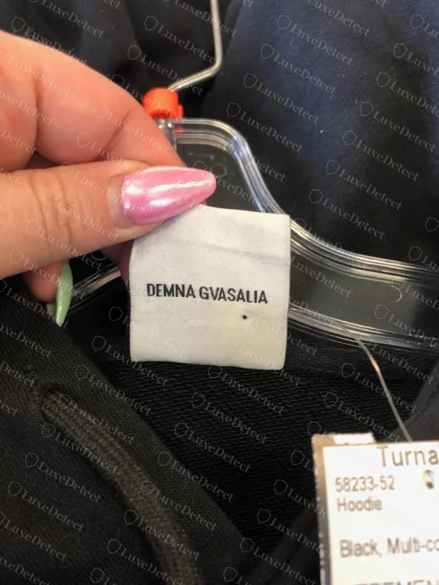 Genuine Vetements white woven label reading DEMNA GVASALIA, held up on a hanger inside the neck of a black hoodie with a retailer tag beside it