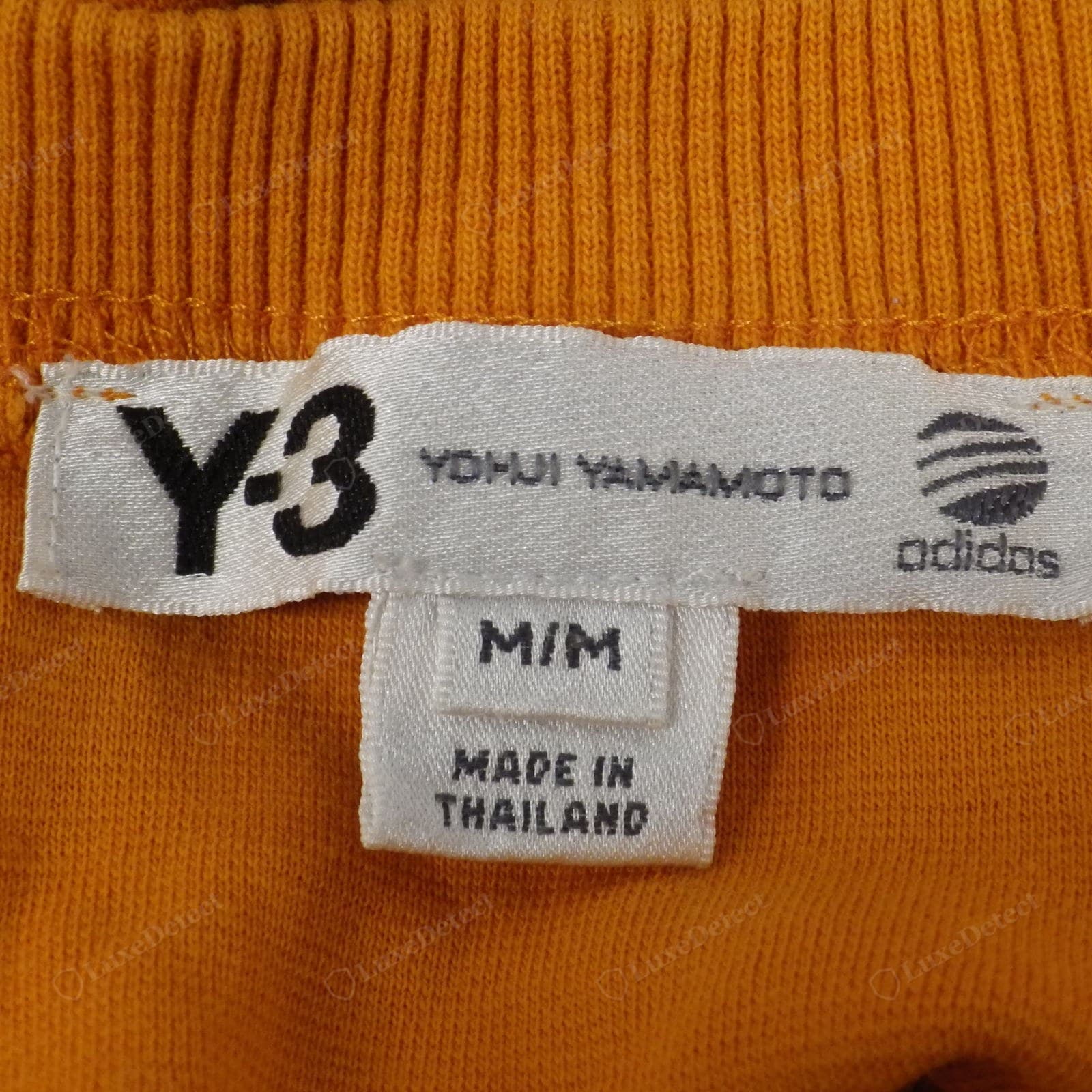 Counterfeit Yohji Yamamoto Y-3 label — the "YOHJI YAMAMOTO" text and the adidas globe logo are blurred and pixelated, the label weave coarse, on a "MADE IN THAILAND" tag