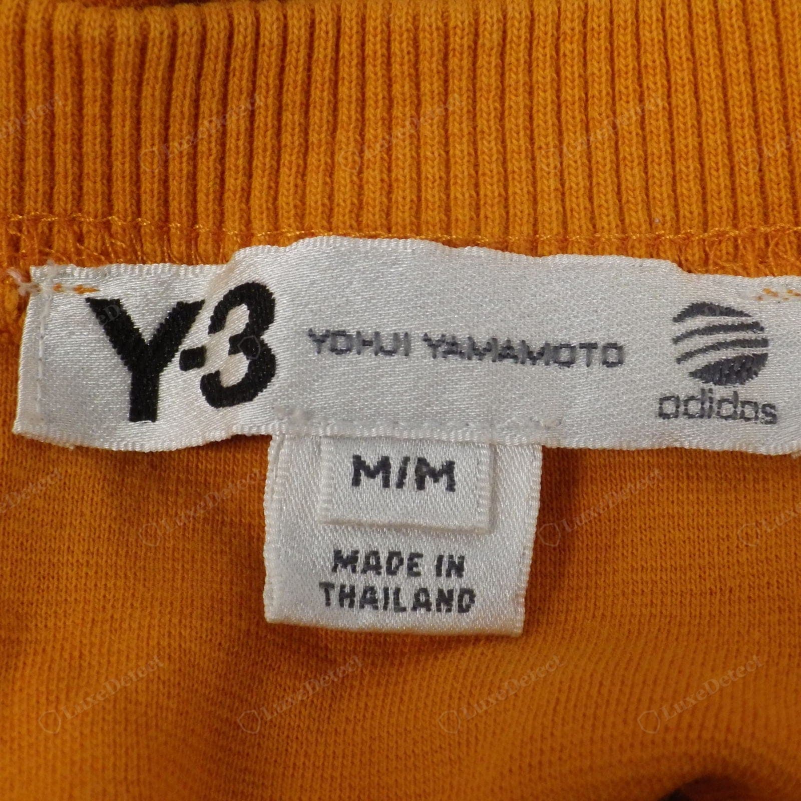 Counterfeit Yohji Yamamoto Y-3 label — the "YOHJI YAMAMOTO" text and the adidas globe logo are blurred and pixelated, the label weave coarse, on a "MADE IN THAILAND" tag