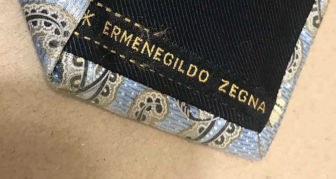 Genuine Ermenegildo Zegna tie label — "ERMENEGILDO ZEGNA" woven in gold thread on black, with the house's small monogram device, sewn into a paisley silk tie