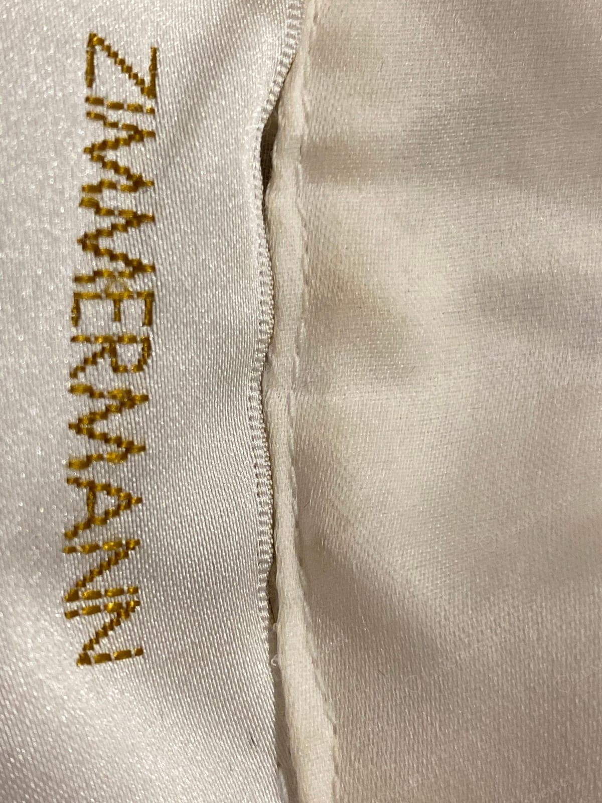 Counterfeit Zimmermann label — "ZIMMERMANN" in coarse brassy gold embroidery, the stitching jagged and the letters lumpy and irregular, a second "M" malformed